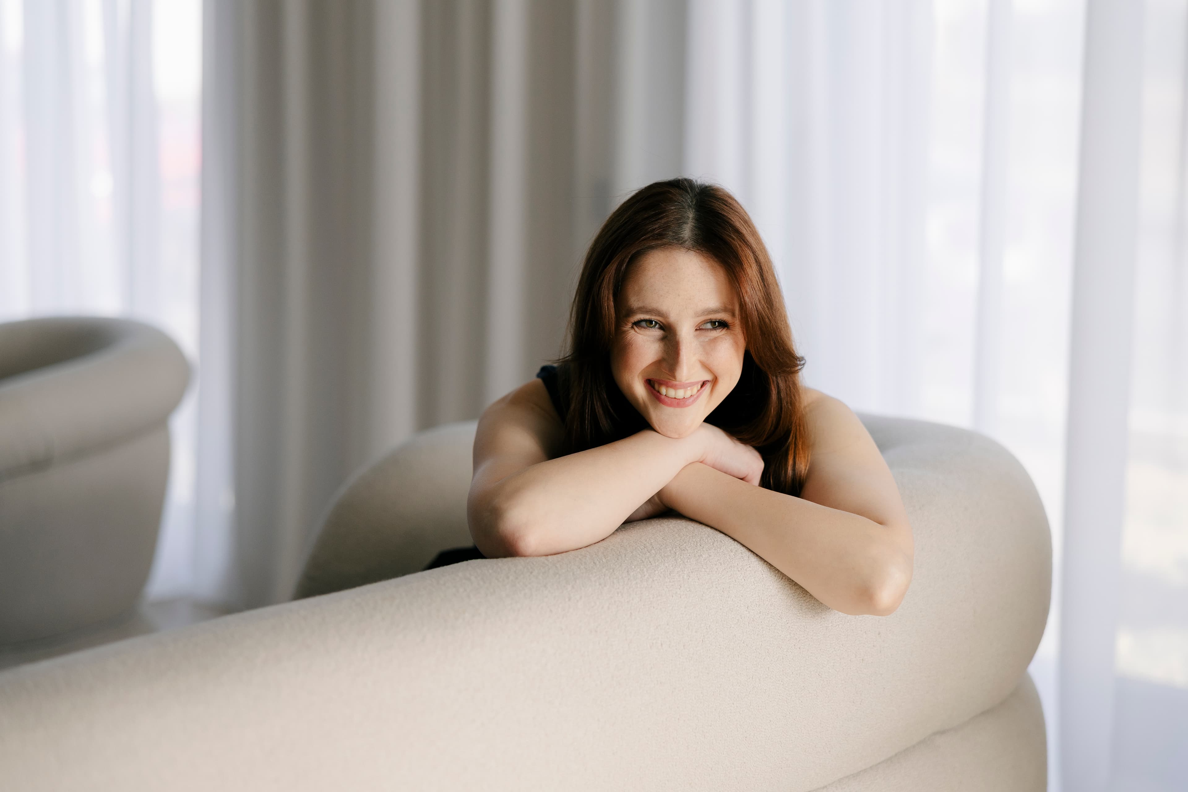 Lorena Răuță, wedding & event planner, zâmbind sprijinită de o canapea crem, în lumină naturală